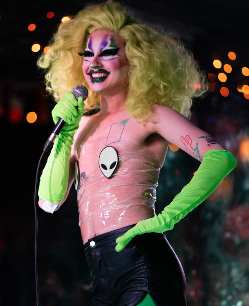 Drag queen with green hair poses on stage with microphone in hand