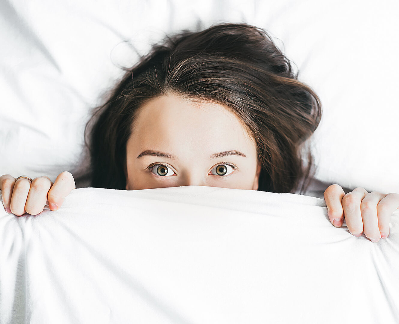 woman lays in bed peeking her eyes out from under the covers