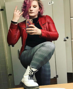 woman in plaid pants and red leather jacket poses in bathroom mirror