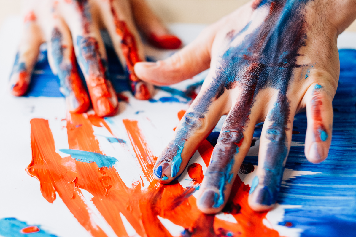 Two hands are covered in paint, resting on white paper also covered in paint.