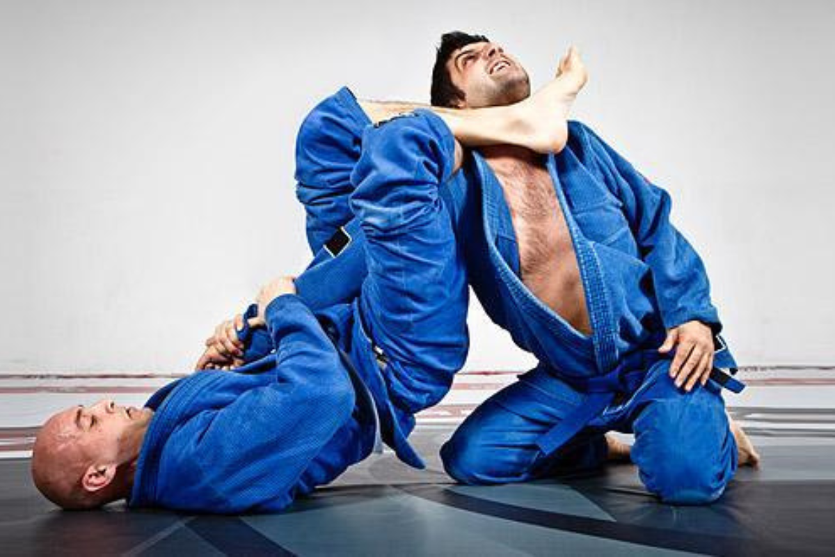 Two men in blue gi's spar on a mat. One mans legs are around the other's neck.