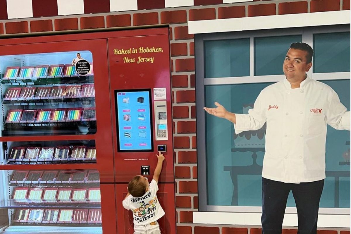 A cutout image of Buddy Valastro from Cake Boss placed beside a cake ATM. A child is seen reaching to press a button on the cake vending machine.