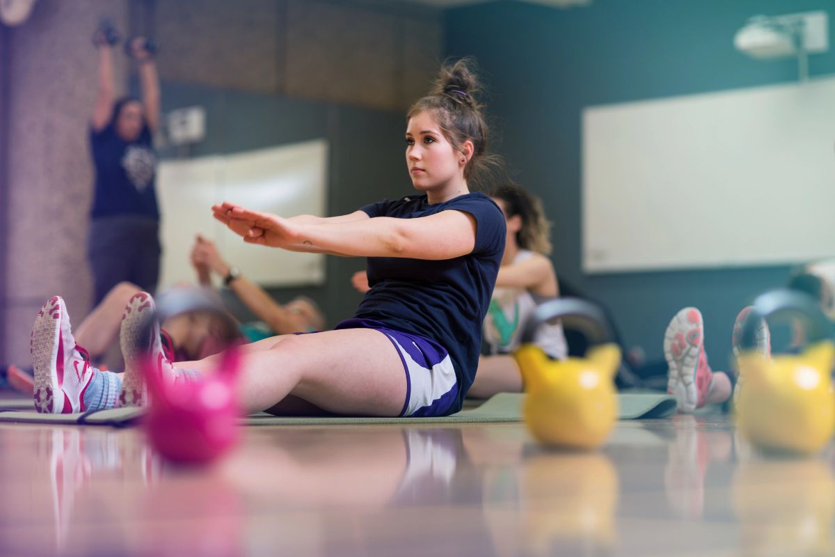 a woman in shorts and a tshirt sits on a gym floor. her legs are extended and her arms are raised in front of her. there are two out of focus kettlebells in front of her.