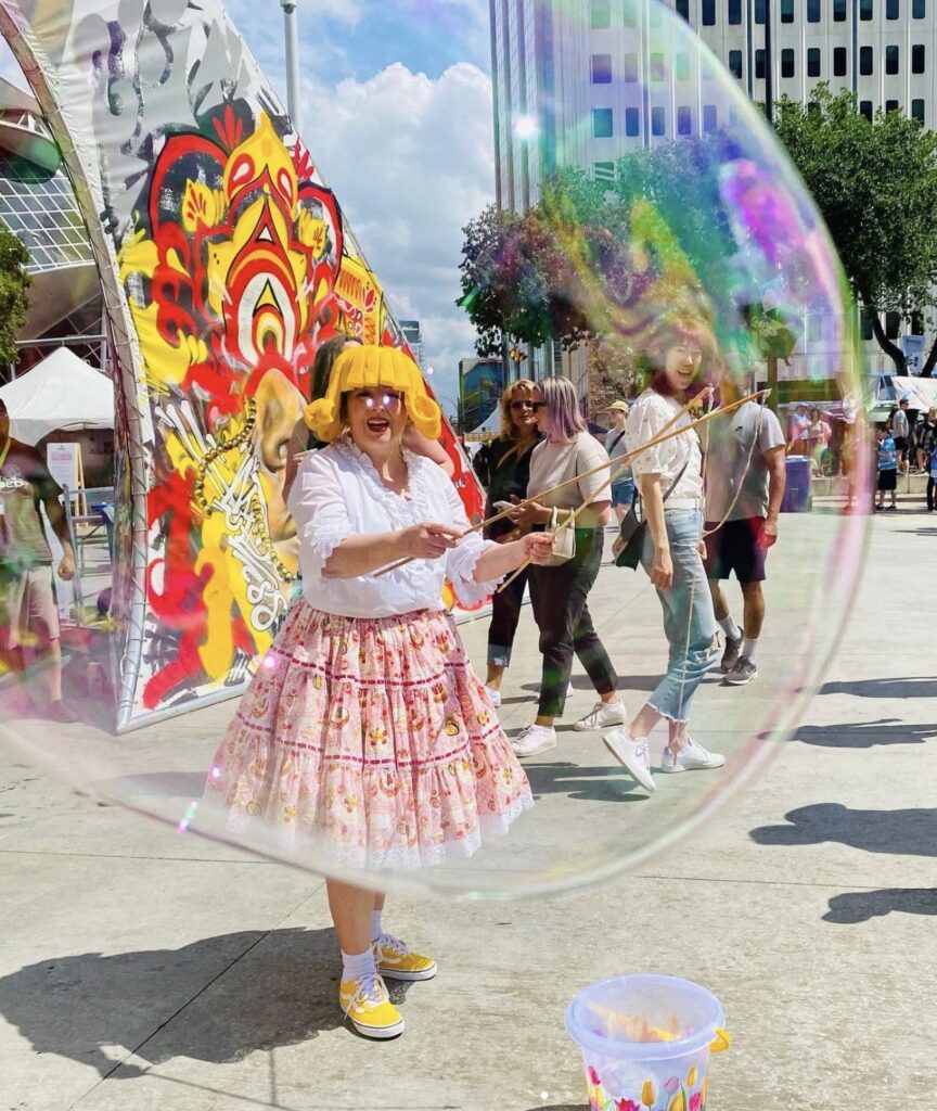 A woman in a yellow wig and colourful skirt blows a giant bubble. The bubble covers three people. 