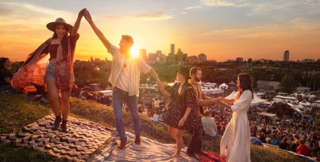 Four people dance on the top of Gallagher Hill. There are tents and crowds of people in the background and the sun is setting. 