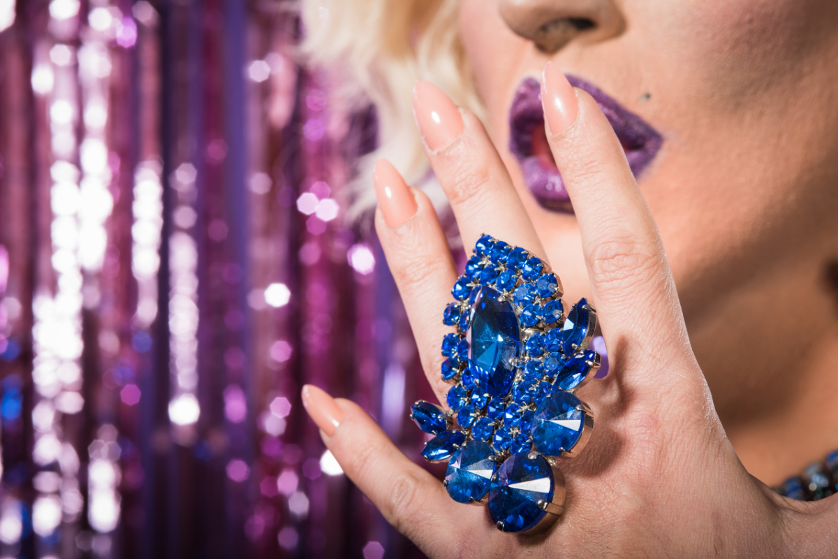 A drag queen is wearing a blue ring with lots of diamonds and holding it up to her face.