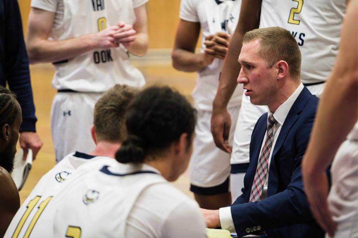 Jordan Baker, coach of the men's ooks basketball team, wears a suit and talks to his team. Players stand and sit around him, all wearing white NAIT Ooks jerseys.