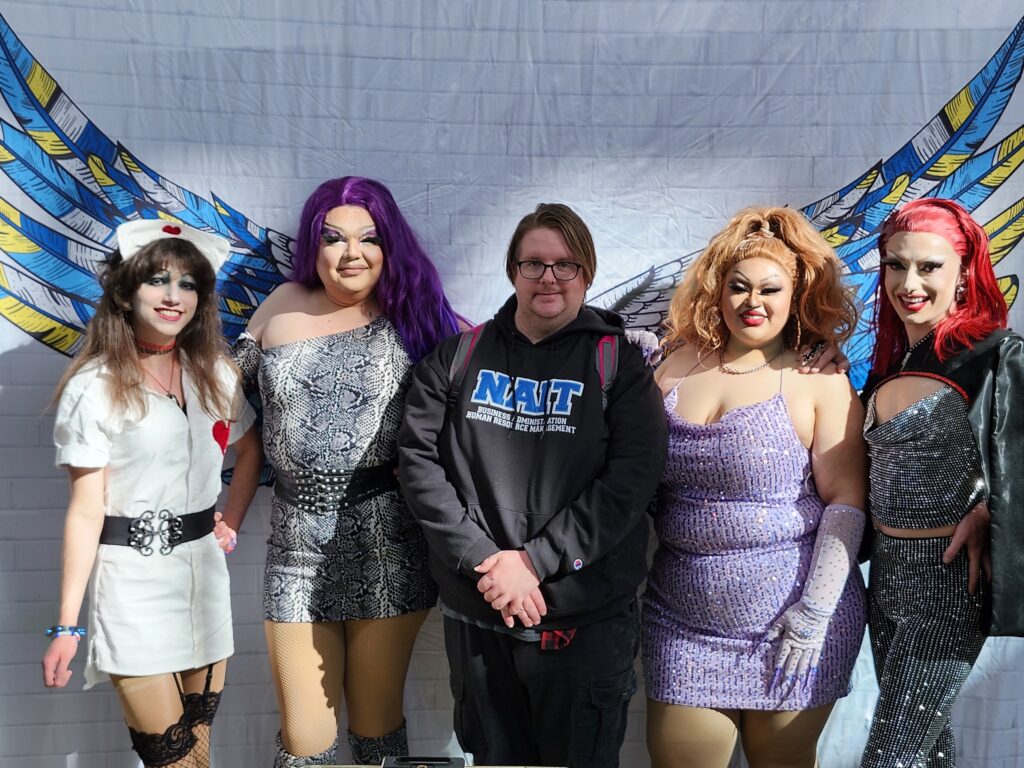 Four drag queens wearing colourful makeup and outfits pose with a student wearing a NAIT HR hoodie 