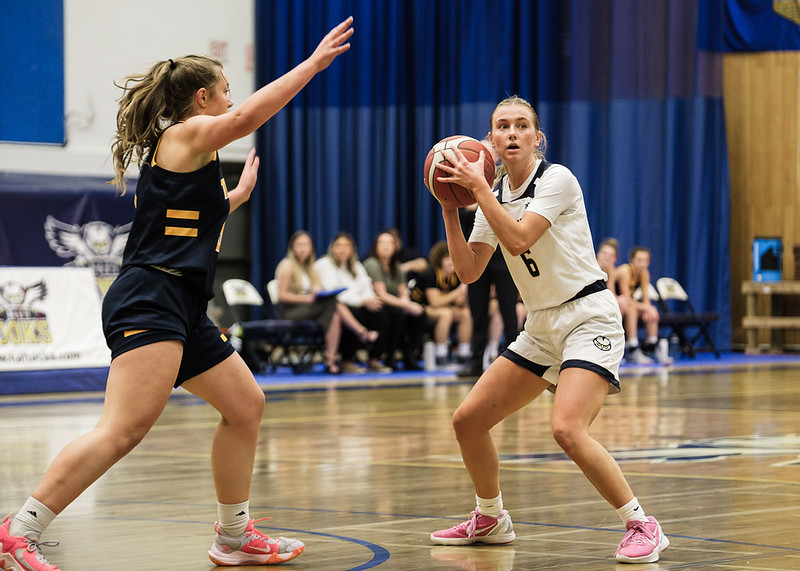 A blonde female basketball payer crouches in a defensive stance. She holds the ball in her hands, and an opponent reaches out to block her upcoming shot. 
