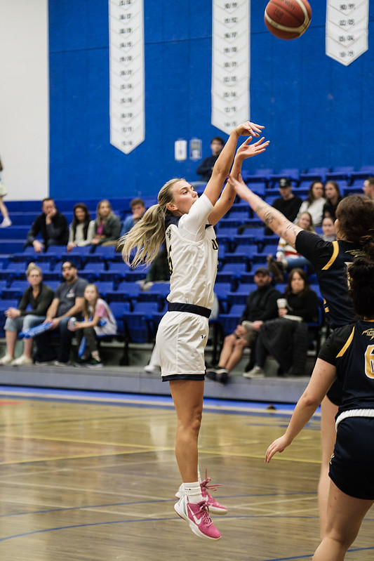 A blonde woman wearing a basketball uniform jumps in the air after shooting the basketball. An opponent is reaching to block her shot. 