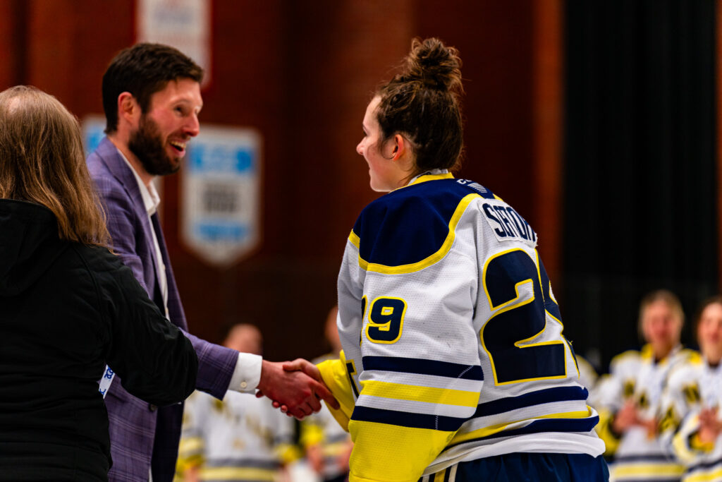 Woman in hockey jersey with hair in a bun shakes hand of man in a suit. Both are smiling. 