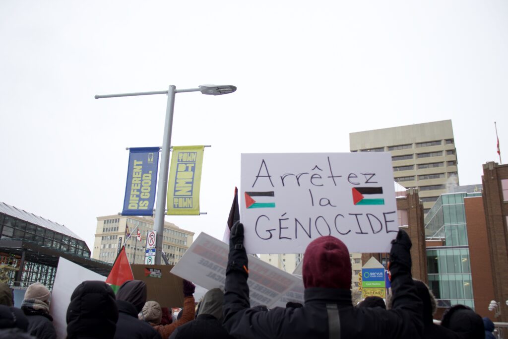 A crowd marches, pictured from behind. Someone holds a sign that says "arretez la genocide" with a handdrawn Palestine flag. 