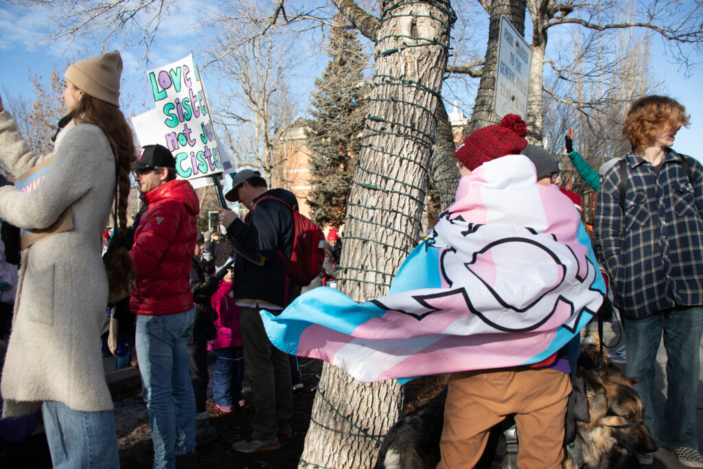 A protestor wears a trans pride flag as a cape. The flag has blue and pink stripes and it billows in the wind.