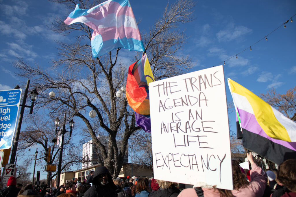A sign reads "the trans agenda is an average life expectancy." the sign is white with black writing. it looks as though it has been handwritten. pride flags billow in the background, and you can see the heads of a large crowd.