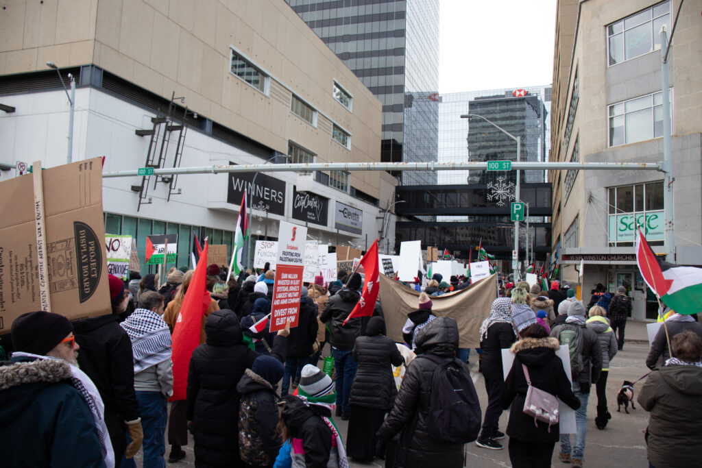 A crowd marches in downtown edmonton. many of them wear red and green clothing. they are marching for palestine. 