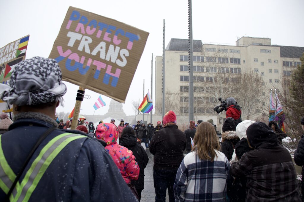 a person in a crowd holds a sign that says "protect trans youth." the letters are pink, blue and white striped. 