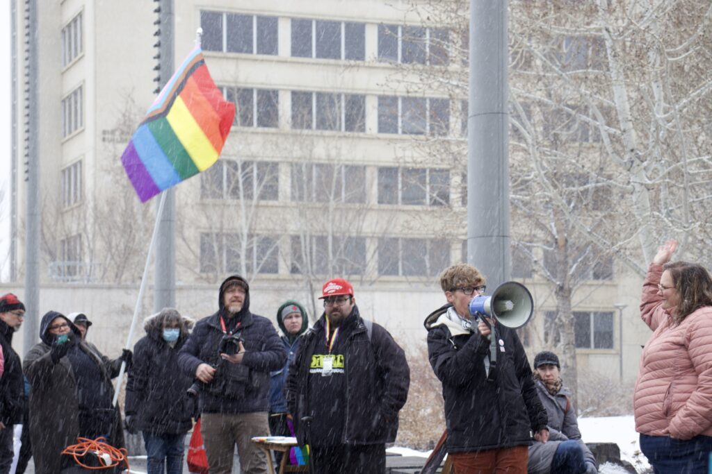 Janis Irwin speaks to a crowd with a megaphone. A person is waving a pride flag in the background. 