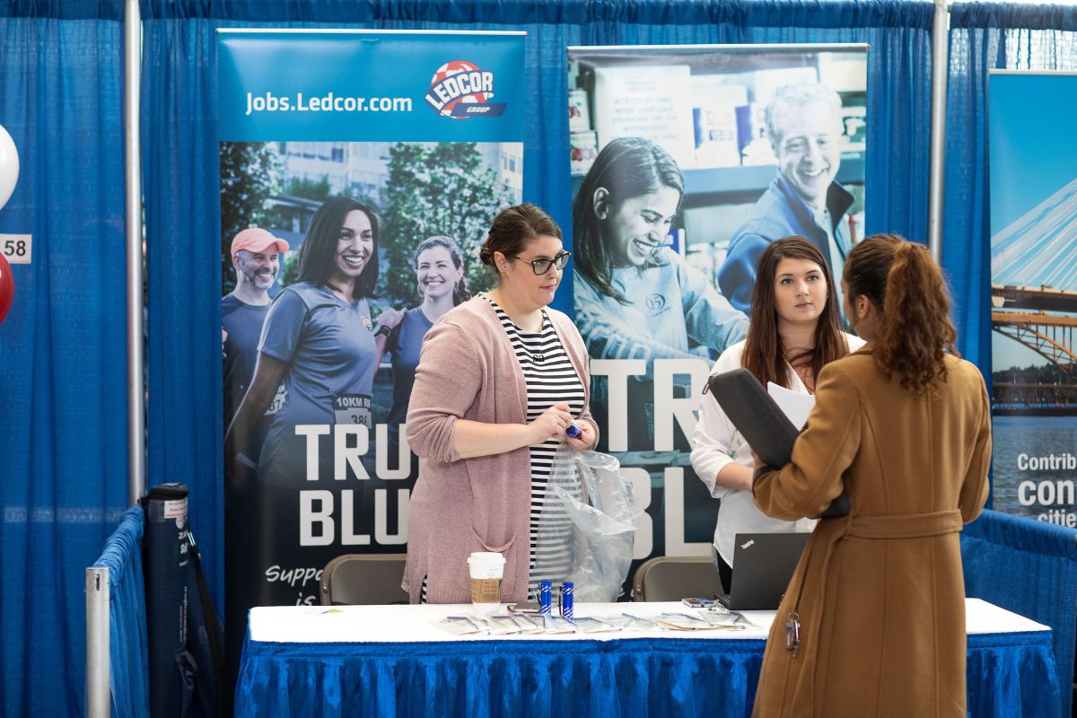 two people stand behind a booth at a hiring fair. a person in a brown sweater stands in front of the table, holding a binder in their arms.