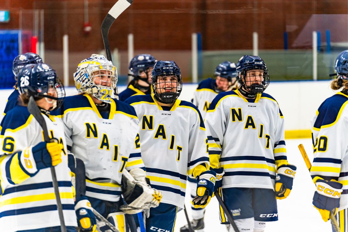 Players on the Ooks hockey team smile in their hockey gear. Their jerseys are mostly white with blue and yellow stripes on the bottom and shoulders. NAIT is written diagonally across the chest.