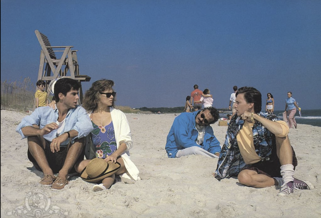 four people sit on a beach. one guy in the back looks like he is asleep. 