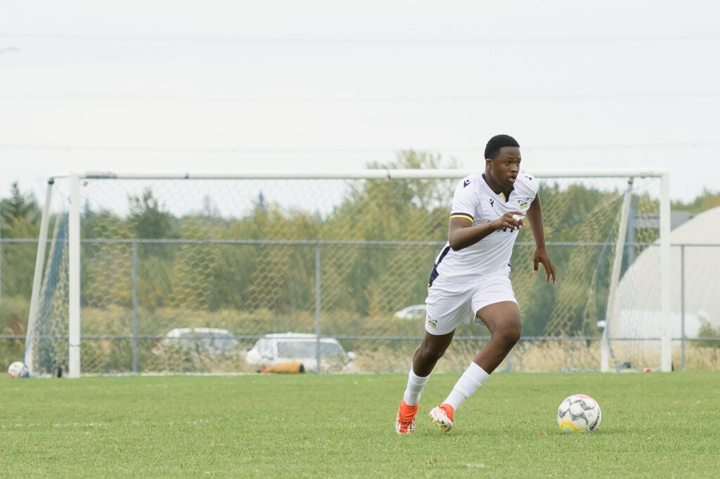 A dark-skinned man in a white soccer uniform runs down a soccer field with the ball at his feet. 