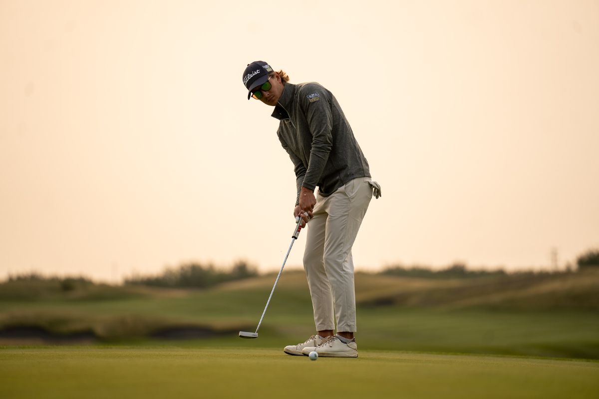 A white man in a blue jacket and beige pants holds a golf club and is poised to make a shot. The sky is a dusky orange behind him.