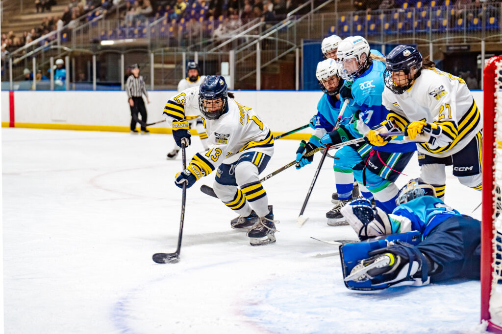 NAIT hockey player makes a shot on the Broncos goalie, who is sprawled on the ice attempting to block. 