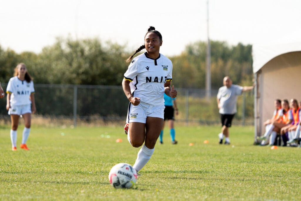A brown-skinned woman with a long braid runs with a soccer ball. She's wearing a white NAIT soccer uniform. 