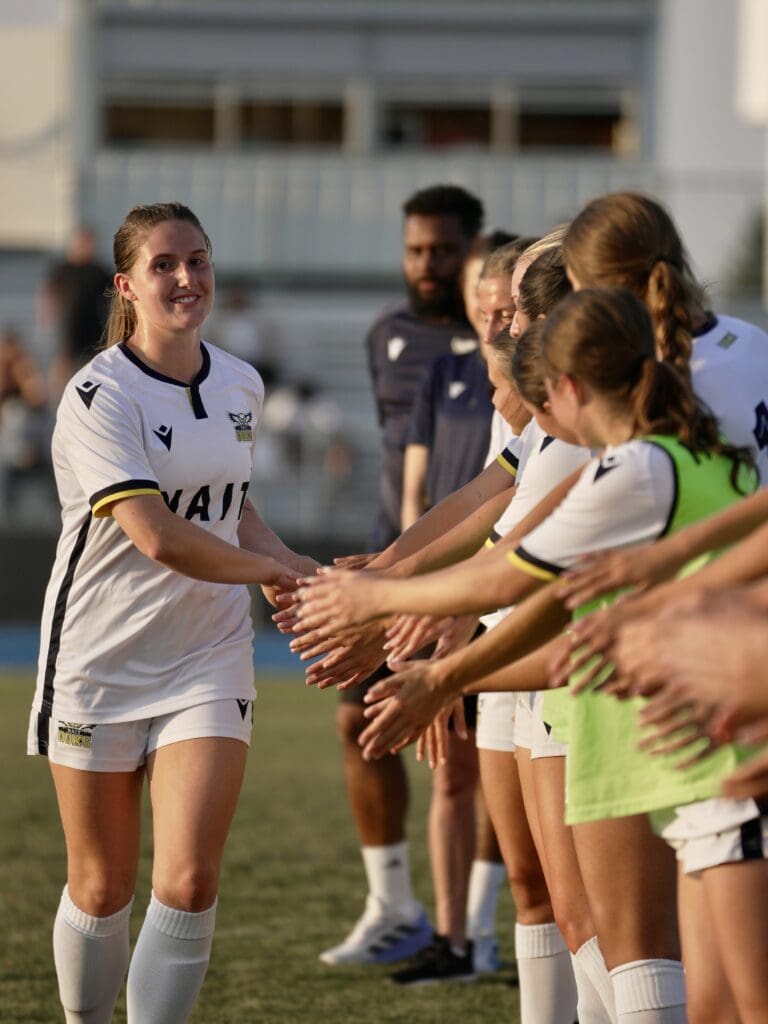 A woman in a white soccer uniform claps the hands of her teammates in line. 