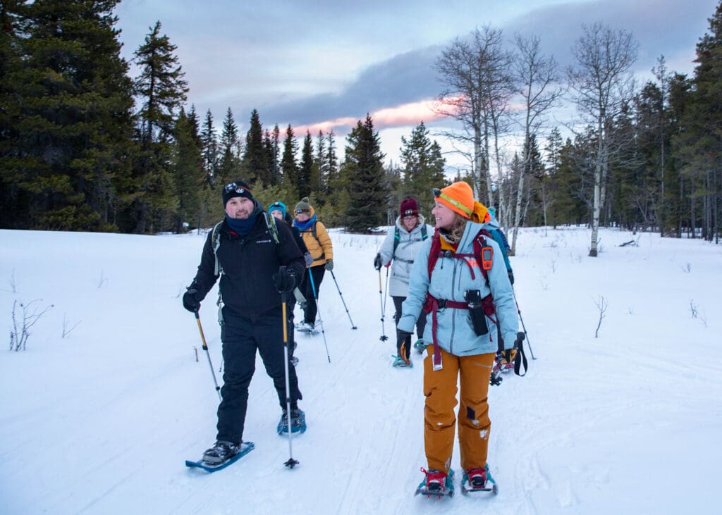 A group of people in winter gear walk on snowshoes along a snowy path. 