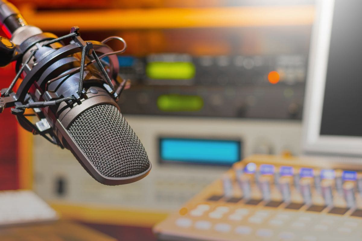Image of inside a radio booth, with a microphone in focus and control panels in the background.