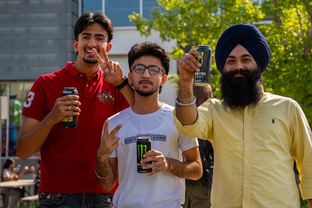 Three men pose for the camera. Two give peace signs, and the other holds up a can of Monster Energy. 