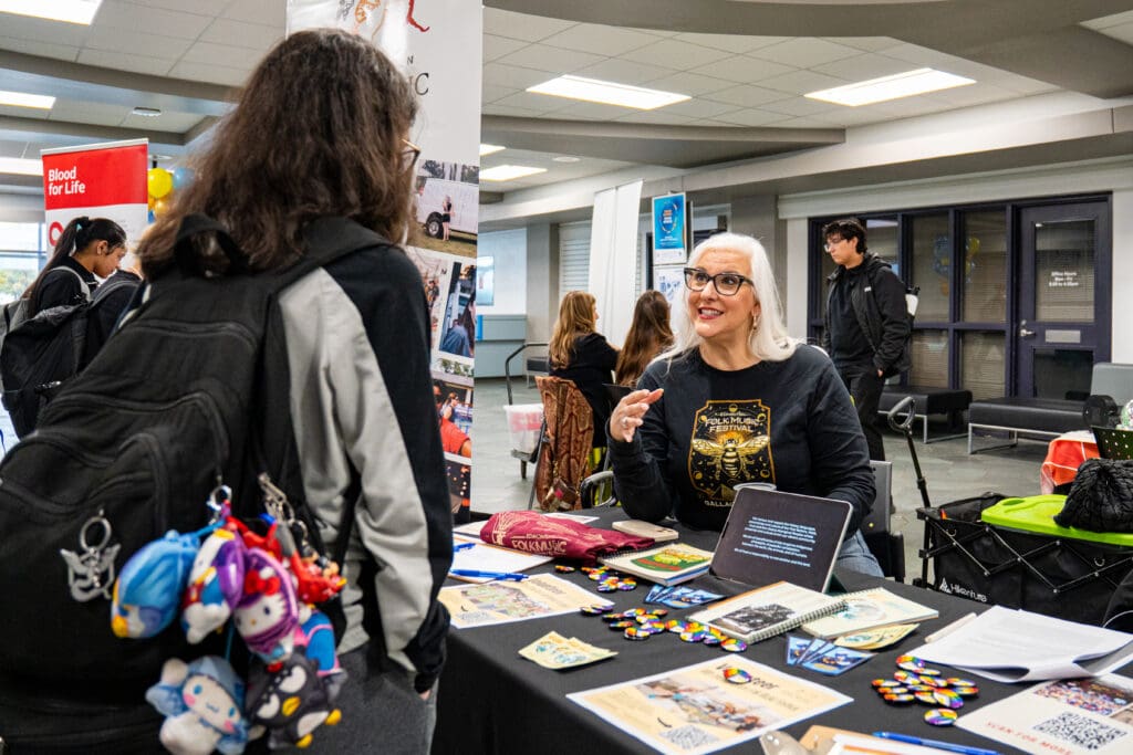 A behind-the-shoulder shot of a young talking to someone at a table. The person in the shot is wearing a sweater that says Folk Music Festival and is smiling.