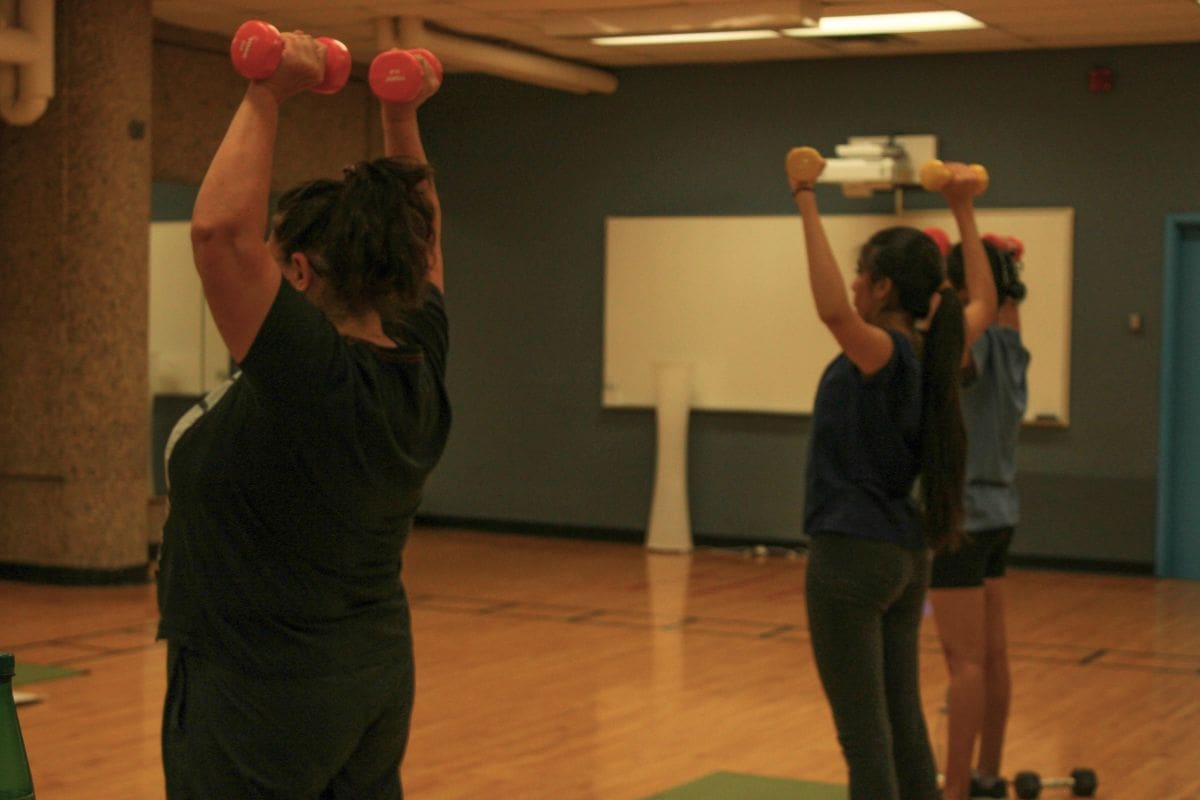 Three women hold dumbbells in the air. The photo is taken from behind.